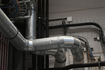 Industrial metal ductwork with bends and joints, part of HVAC system, mounted near ceiling with pipes and cable tray in background.