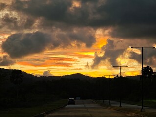 Orange light bathes the summer city landscape and asphalt highway, with a silhouette of a car traveling toward the evening horizon under dramatic dusk clouds