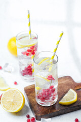 Two tall glasses with lemon red currant water and straws on wooden board with ice cubes