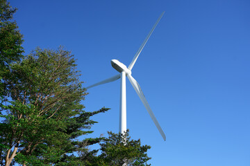 A giant wind turbine stands powerfully against a vibrant blue sky and green trees. The scene symbolizes the harmony of nature and technology, representing clean energy and a sustainable society.