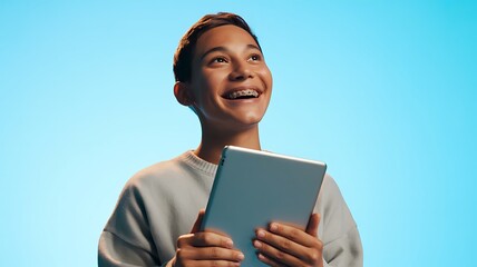 Vibrant studio portrait of a bright, happy teenager with braces holding a tablet and looking up with optimism and excitement for the future.

