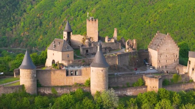 bourscheid castle ch&acirc;teau de green cover hills walls towers flags atop buildings visitor vehicles parked nearby to left around right up close circling drone shot 