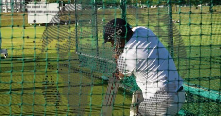 Practicing batsman wearing helmet, gloves and pads swinging bat by stumps inside green net cage