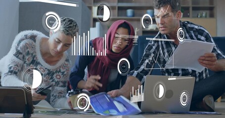 Collaborating team analyzing charts around table in lounge with laptop tablet smartphone coffee cup