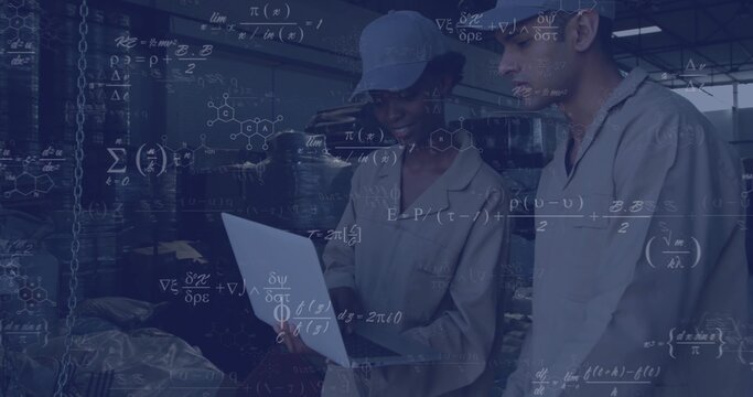 Analyzing data, technicians in coveralls monitoring laptop on plant floor, with drums, shelving