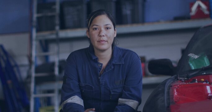 Kneeling female mechanic wearing navy coveralls inspecting red car engine in shop, with shelves