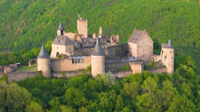 bourscheid castle ch&acirc;teau de green cover hills walls towers flags visitor vehicles old historical site surrounded by to left around right up close drone panning 