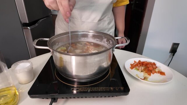 A woman in an apron cooks soup on an induction hob, stirring the soup with a spoon