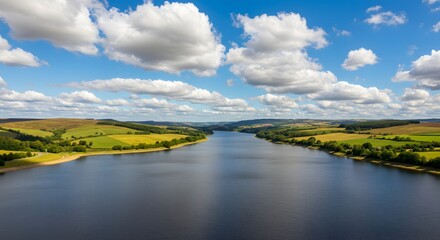 Aerial view of a calm lake surrounded by rolling hills, fields, and a bright, cloudy sky