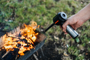 Close-up of a person using a portable air blower to ignite a fire in a metal firepit. Fire safety, controlled burning, outdoor cooking, camping equipment
