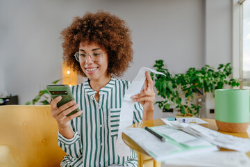 Afro woman using smart phone and calculating financial bills at home
