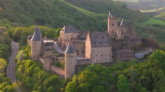 bourscheid castle ch&acirc;teau de green valley cover hills behind walls black domed towers road beside historic site trees to left around right drone panning shot 
