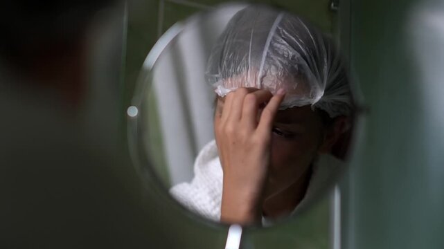 A girl looks at herself in a round mirror in a hotel bathroom and adjusts the clear shower cap on her hair. The teenager is performing her morning or evening body care routine.