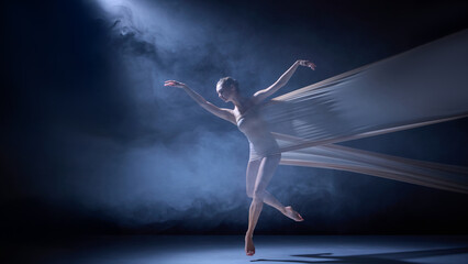 Ballet dancer in fabric performing elegant leap under blue haze light. Concept of choreography, dynamic stage performance, and physical artistry in dance production.