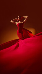 Fototapeta premium Female model in red dress posing under warm studio light with flowing fabric. Concept of fashion style, elegance, color harmony for editorial, commercial, and design projects.
