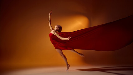 Ballet dancer in red fabric leaping under warm orange spotlight in studio. Concept of movement,...