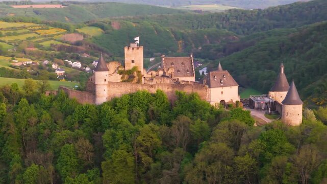bourscheid castle ch&acirc;teau de green valley cover hills behind walls black domed towers flag top foggy weather colorful trees to left near right drone panning shot 