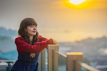 An elegant takes a deep breath, feeling happy, confident while enjoying her free leisure time on the rooftop of luxury hotel. The sun over clouds in the sea. Evening light with beautiful orange sky.