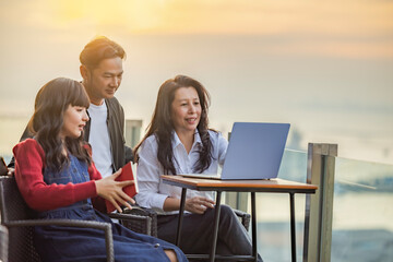 Small diverse business team of three colleagues meeting at laptop, watching online startup presentation, website content, talking on rooftop near the sea. Colours of sunset.Skyscape nature composition