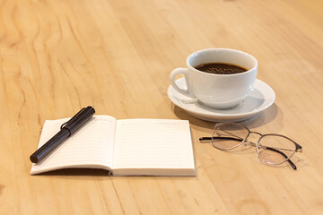 Warm coffee sits beside an open notebook and a pen on a wooden table in a cozy setting