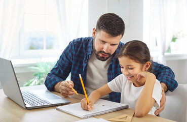 Father and daughter learning homework together. Dad guides a smiling child at a desk, with laptop...