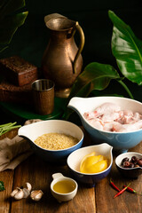 A warm, rustic kitchen still life featuring raw chicken, rice, lemon slices, olives, garlic, olive oil, and a copper pitcher among green leaves and wooden textures for a cozy culinary scene.