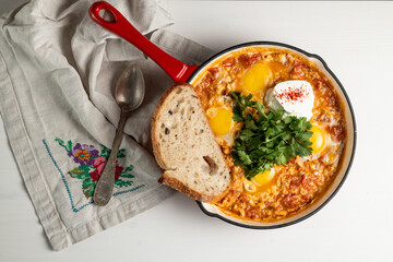 Delicious shakshuka, a hearty breakfast or brunch, featuring eggs poached in a vibrant tomato sauce, garnished with fresh herbs and sour cream, served with rustic bread in a red cast-iron skillet.