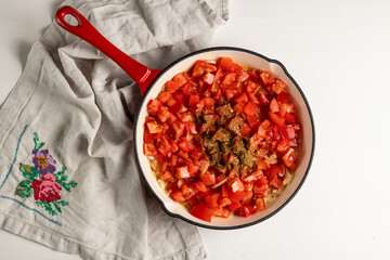 Skillet with diced tomatoes, onions, and spices on embroidered linen. Food preparation from above.