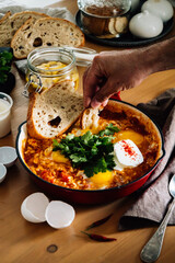 Vibrant shakshuka with poached eggs, fresh cilantro, and crusty bread. A hand dips into the hearty dish on a rustic wooden table, ideal for a delicious breakfast or brunch.