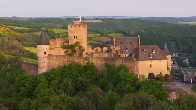 bourscheid castle ch&acirc;teau de green valley cover hills behind walls towers windmills hilltops cloudy sky town below from toward afar forward drone shot 