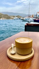 Vertical photo. Bright yellow ceramic coffee cup with sea, port, and colorful Greek town in background