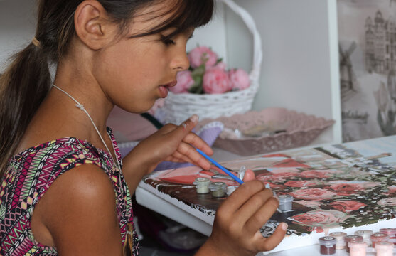 A girl paints a picture using paints and a brush. Paint by numbers. A fragment of the canvas and paint containers are visible.
