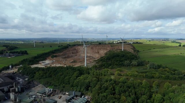 Aerial view of wind turbines standing tall near a quarry, juxtaposed against lush green fields and a dense forest, Matlock, England, United Kingdom.