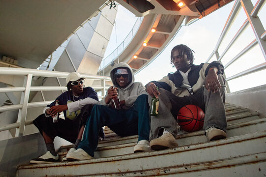 Three Black men sitting on outdoor stairs, holding drinks and basketball, wearing casual streetwear, looking relaxed and engaged in conversation under modern architectural structure