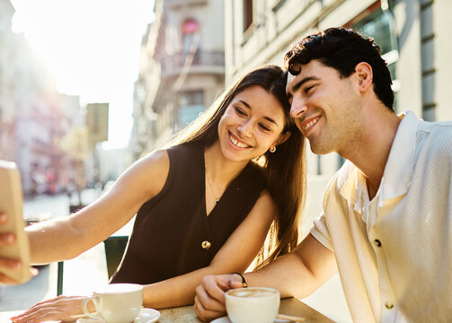 Young couple enjoying conversation and coffee together and taking a selfie with a smartphone at an outdoor urban cafe table