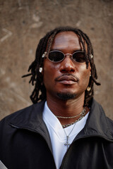 Fototapeta premium Portrait of young adult Black man wearing sunglasses and jewelry looking directly at camera standing against textured wall background, displaying confident facial expression