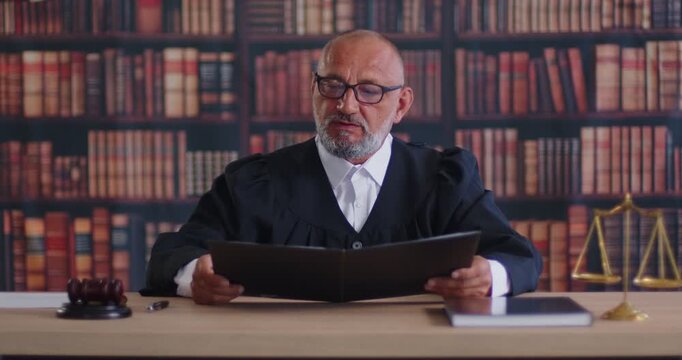 Judge reading criminal case document in courtroom. Senior magistrate holding folder to review court order, man in lawyers robe sitting at wooden desk with gavel and balance scales, law books behind