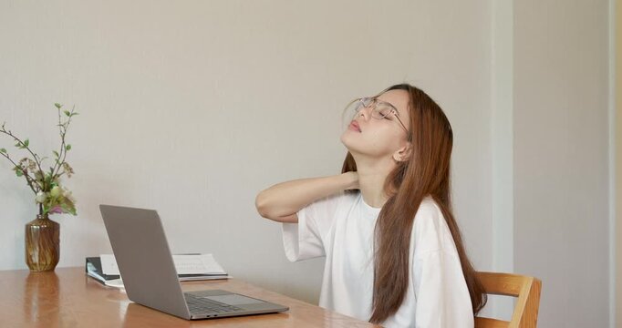 Asian adult woman rubs neck while sitting at laptop during home work session, suffering from stiffness and muscle strain caused by long working hours and uncomfortable chair in quiet room