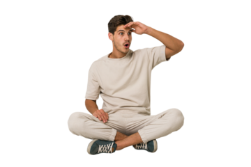 Young caucasian man sitting on the floor isolated on white background looking far away keeping hand on forehead.