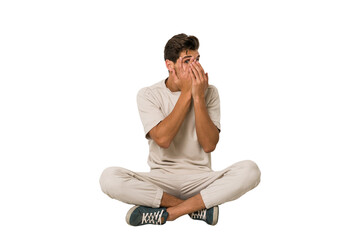 Young caucasian man sitting on the floor isolated on white background blink through fingers...