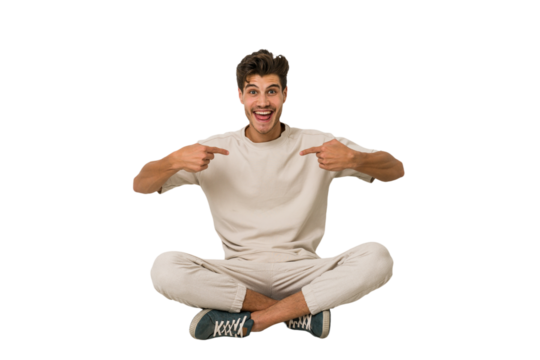 Young caucasian man sitting on the floor isolated on white background surprised pointing with finger, smiling broadly.
