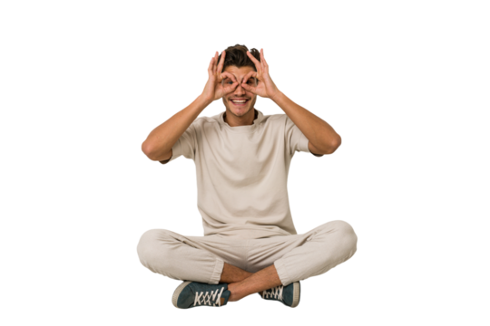 Young caucasian man sitting on the floor isolated on white background showing okay sign over eyes