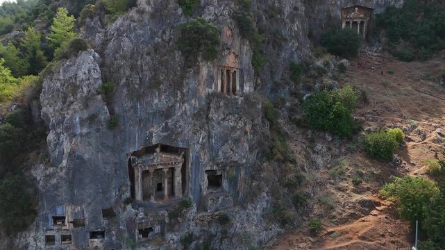 Aerial view of the ancient Lycian rock tombs of Telmessos in Fethiye, Turkey, carved into a steep mountain cliff surrounded by trees and rugged landscape.