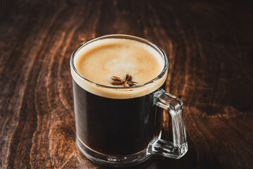 Glass mug with dark beer or coffee topped with creamy foam and star anise, placed on rustic wooden surface. Warm, aromatic, seasonal drink concept.