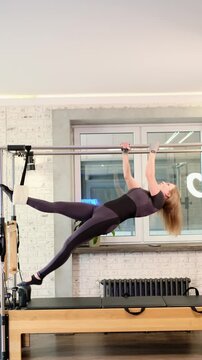 A woman performs a Pilates V-sit exercise on a reformer tower, holding the bar firmly while keeping her legs lifted. The frame from real workout footage illustrates balance, strength, and control.