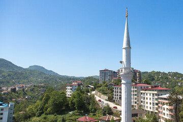 Obraz premium View of Rize city in Turkey with a white mosque minaret, modern residential buildings, green hills, and clear blue sky, representing culture, religion, architecture, and travel concepts