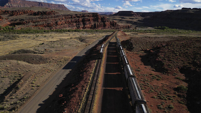 Aerial view of a long train stretching across the arid landscape beneath towering red rock formations, a stark contrast against the vast desert, Moab, Utah, United States.
