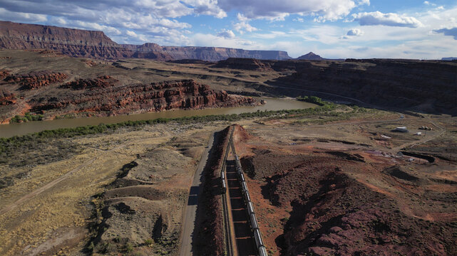 Aerial view of a long train snaking through the arid landscape near the Colorado River, framed by towering red rock formations under a vast sky, Moab, Utah, United States. - Powered by Adobe