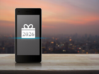 Gift box happy new year 2026 icon on modern smart mobile phone screen on wooden table over blur of cityscape on warm light sundown, Business happy new year 2026 shop online concept