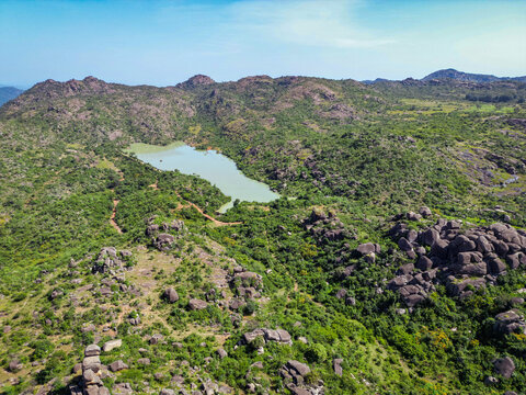 Aerial view of rugged, rocky outcrops interspersed with lush green vegetation surrounding a serene lake, Shere Hills, Plateau, Nigeria.